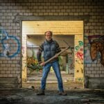 An old man standing holding a pickaxe, walls with graffiti at his back