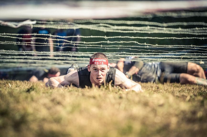 a man with a head band with printed “Spartan 9678”, crawling on the ground, an obstacle course racing